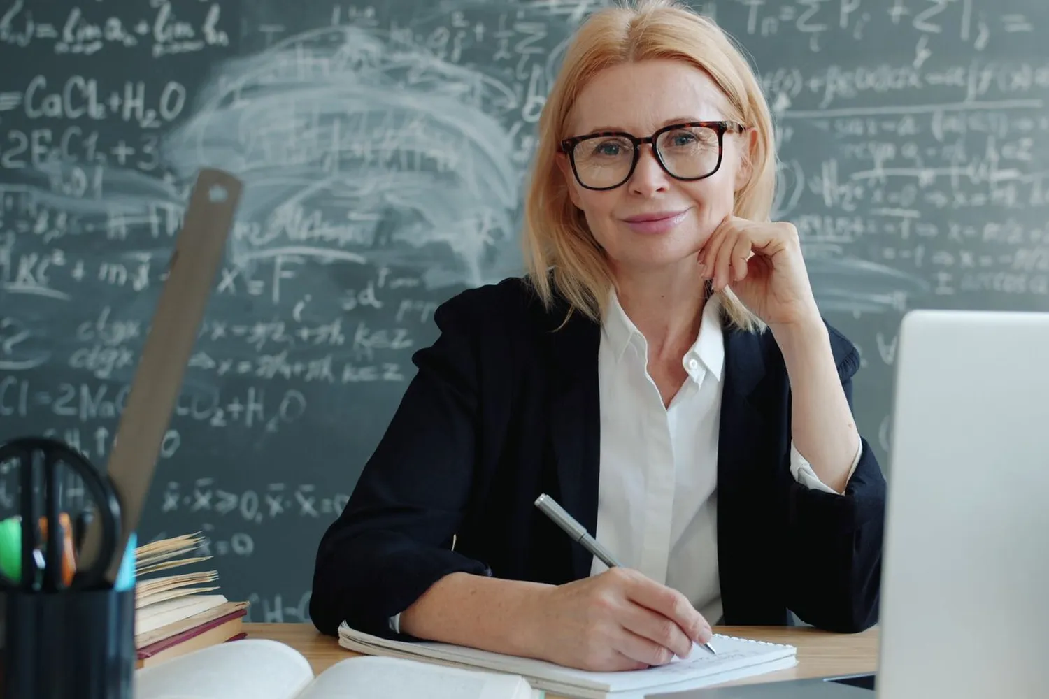 A teacher at her desk with a laptop and a notebook, smiling, in front of a chalkboard